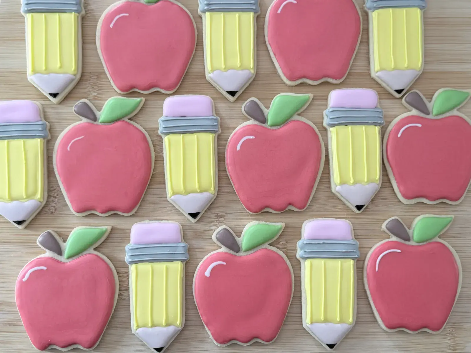 Back-to-school sugar cookies shaped like apples and pencils, iced in pink, yellow, and gray, arranged in neat rows on a wood board.