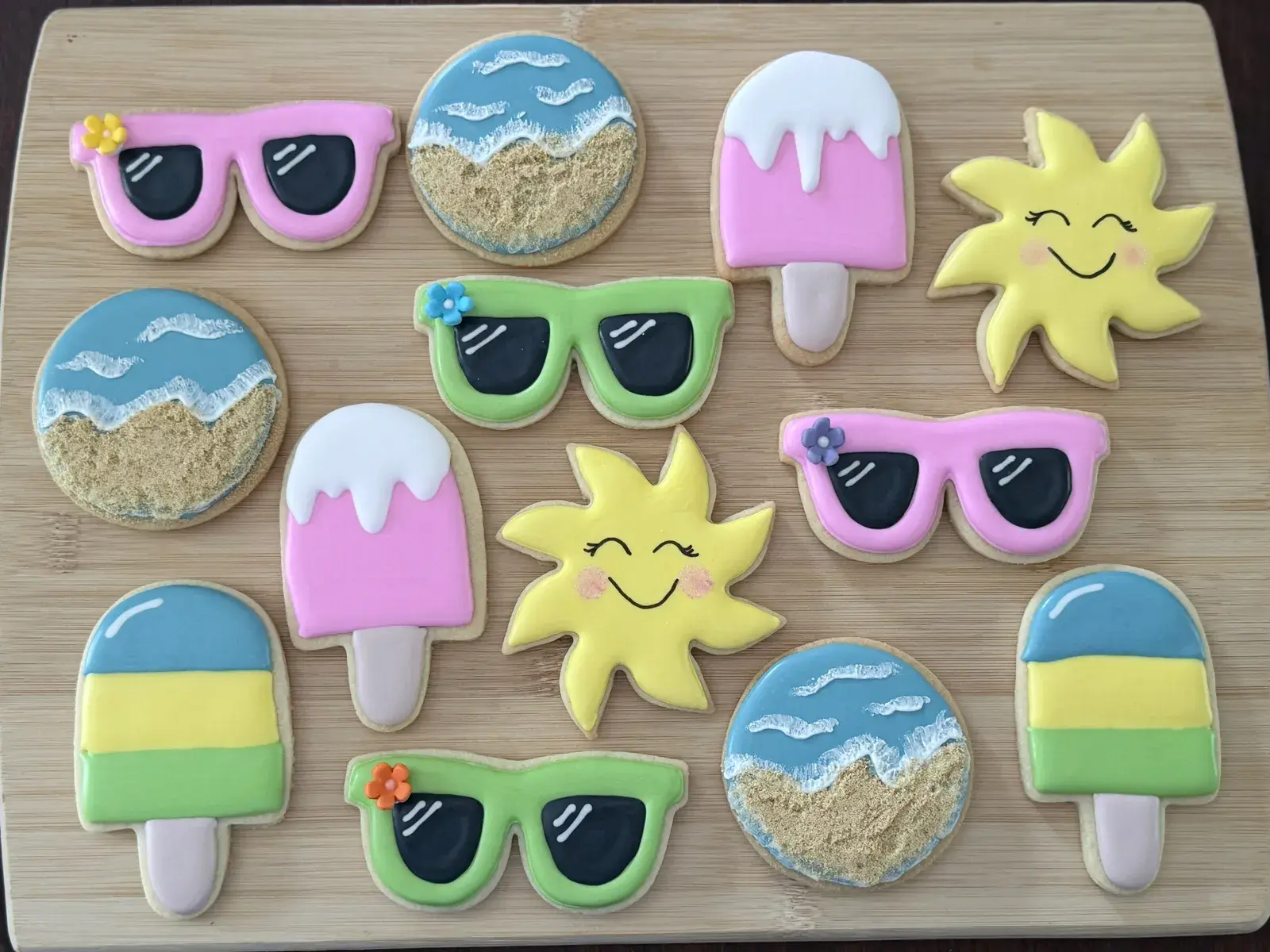 Assorted summer-themed sugar cookies: pink and green sunglasses with tiny flower accents, a smiling yellow sun, a pink-and-white striped popsicle, and a sandy beach scene with blue waves.
