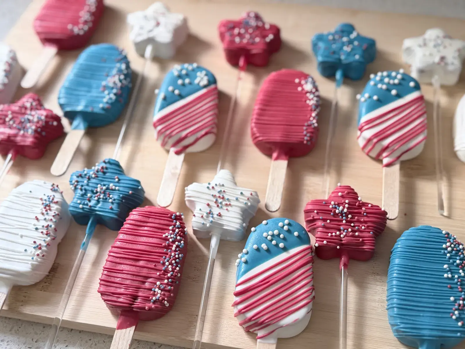 Red, white, and blue chocolate-dipped cakesicles and star-shaped pops on wooden sticks, striped with piped icing and nonpareil sprinkles for a patriotic dessert spread.