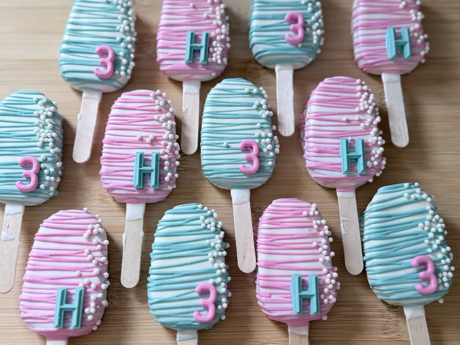 A closer view of the pink-and-teal striped H and 3 cakesicles, showing the fondant letters and numerals up close among the piped stripes.