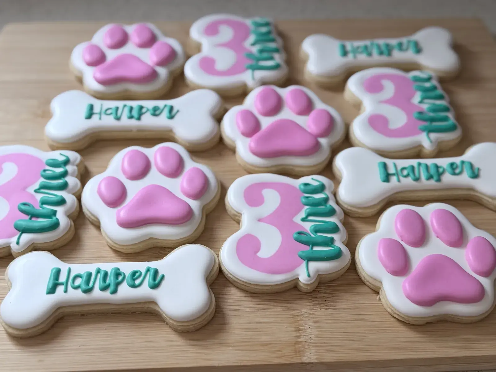 A puppy-themed cookie set filling a wood board: white cookies iced with bright pink paw prints, bone-shaped cookies lettered Harper in teal script, and pink number 3 cookies with the word three piped beside them in teal.